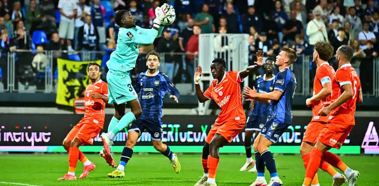 Simon | AllezPaillade.com Soccer goalkeeper in teal leaps to catch the ball as players in orange and blue contest in the box with spectators watching.