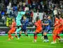 Soccer goalkeeper in teal leaps to catch the ball as players in orange and blue contest in the box with spectators watching.
