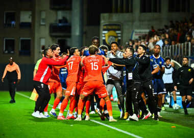 Soccer players in red jerseys celebrate in a tight huddle on the pitch amid teammates and coaches.