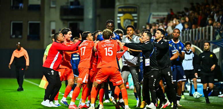 USL Dunkerque v Montpellier HSC - Ligue 2 BKT 20262027 | AllezPaillade.com Soccer players in red jerseys celebrate in a tight huddle on the pitch amid teammates and coaches.