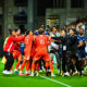 Soccer players in red jerseys celebrate in a tight huddle on the pitch amid teammates and coaches.