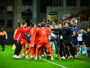 Soccer players in red jerseys celebrate in a tight huddle on the pitch amid teammates and coaches.