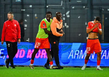 Soccer players on a field; two in orange with a green bib hug a man in orange while another player covers his face with his shirt.
