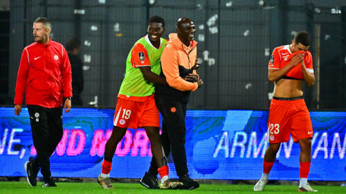 Soccer players on a field; two in orange with a green bib hug a man in orange while another player covers his face with his shirt.