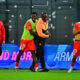 Soccer players on a field; two in orange with a green bib hug a man in orange while another player covers his face with his shirt.