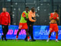 Soccer players on a field; two in orange with a green bib hug a man in orange while another player covers his face with his shirt.
