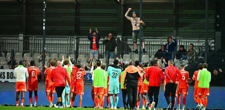 Joie | AllezPaillade.com Soccer players in red celebrate on the field as fans cheer from the stands after a match.