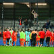 Soccer players in red celebrate on the field as fans cheer from the stands after a match.