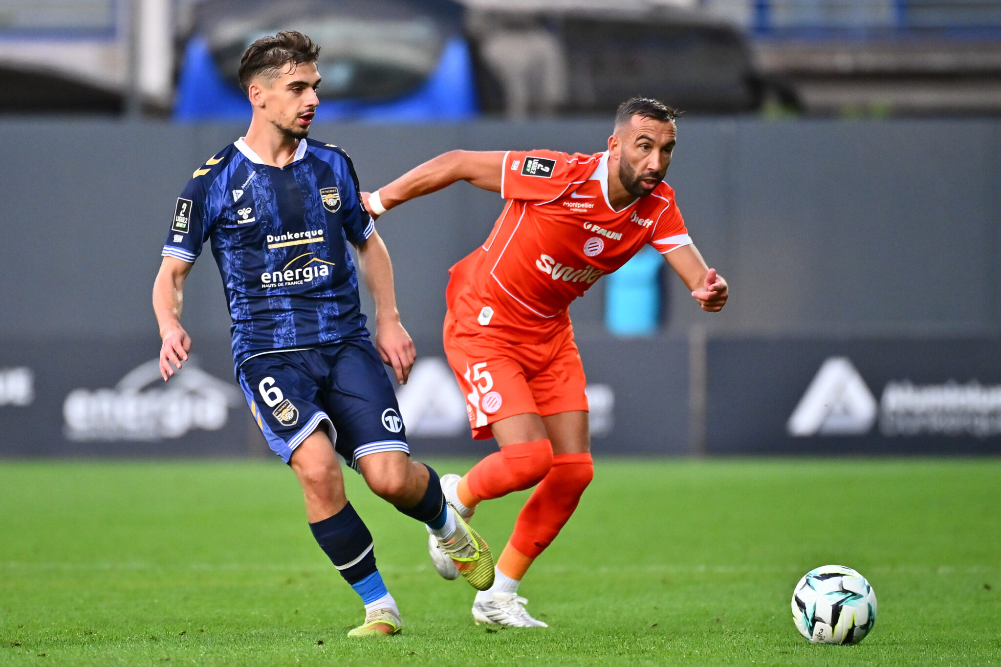 Tardieu USL Dunkerque v Montpellier HSC - Ligue 2 BKT 20262027 | AllezPaillade.com Two soccer players chase the ball on a green field, one in a blue kit and one in orange, during a match.