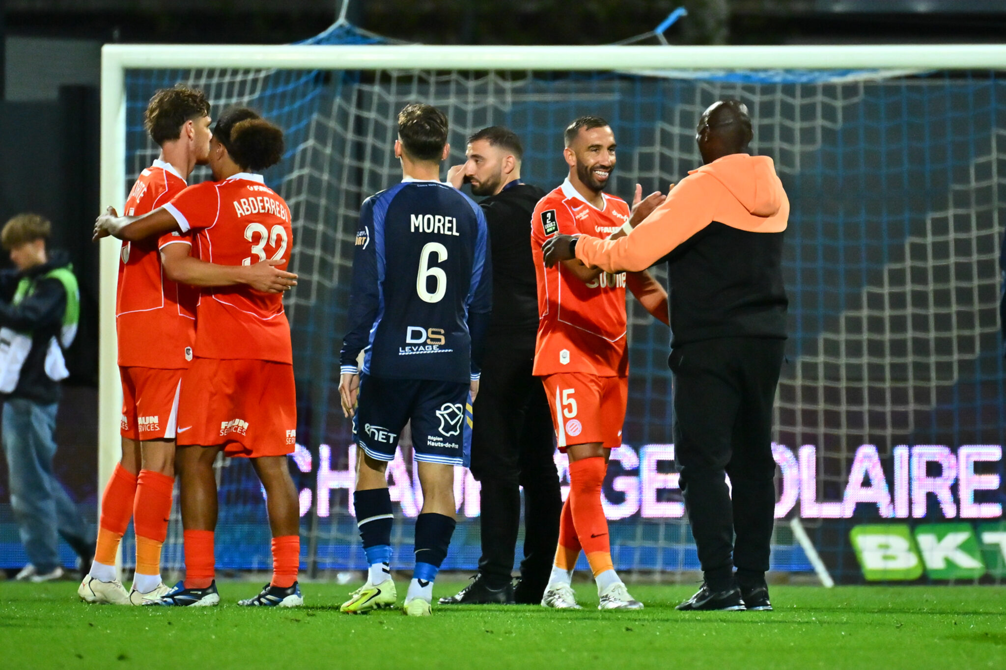 Tardieu Camara USL Dunkerque v Montpellier HSC - Ligue 2 BKT 20262027 | AllezPaillade.com Soccer players in red celebrate in a hug near the goal on a grassy pitch with the net behind them.