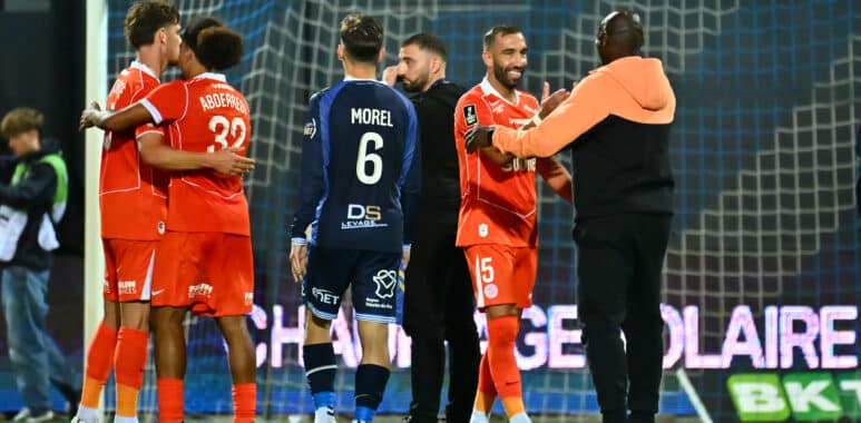 Tardieu Camara USL Dunkerque v Montpellier HSC - Ligue 2 BKT 20262027 | AllezPaillade.com Soccer players in red celebrate in a hug near the goal on a grassy pitch with the net behind them.