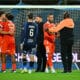Soccer players in red celebrate in a hug near the goal on a grassy pitch with the net behind them.