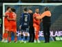 Soccer players in red celebrate in a hug near the goal on a grassy pitch with the net behind them.