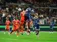 Soccer players in orange and blue jump for a ball in midair as a crowd watches from stands in a packed stadium.