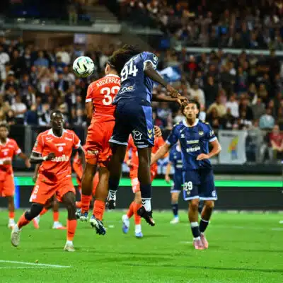Soccer players in orange and blue jump for a ball in midair as a crowd watches from stands in a packed stadium.