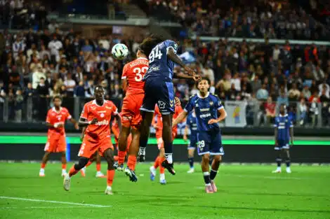 Soccer players in orange and blue jump for a ball in midair as a crowd watches from stands in a packed stadium.