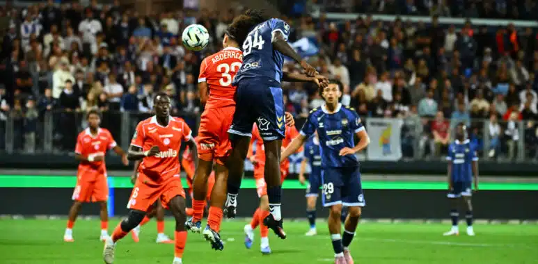 Soccer players in orange and blue jump for a ball in midair as a crowd watches from stands in a packed stadium.