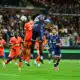 Soccer players in orange and blue jump for a ball in midair as a crowd watches from stands in a packed stadium.