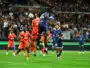 Soccer players in orange and blue jump for a ball in midair as a crowd watches from stands in a packed stadium.
