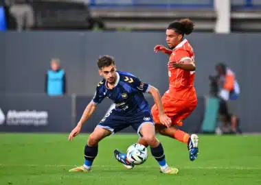 Two soccer players chase the ball during a match, one in blue and one in orange, on a green field.