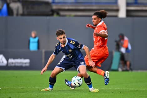 Two soccer players chase the ball during a match, one in blue and one in orange, on a green field.