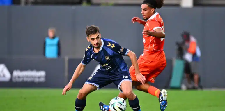 USL Dunkerque v Montpellier HSC - Ligue 2 BKT 20262027 | AllezPaillade.com Two soccer players chase the ball during a match, one in blue and one in orange, on a green field.