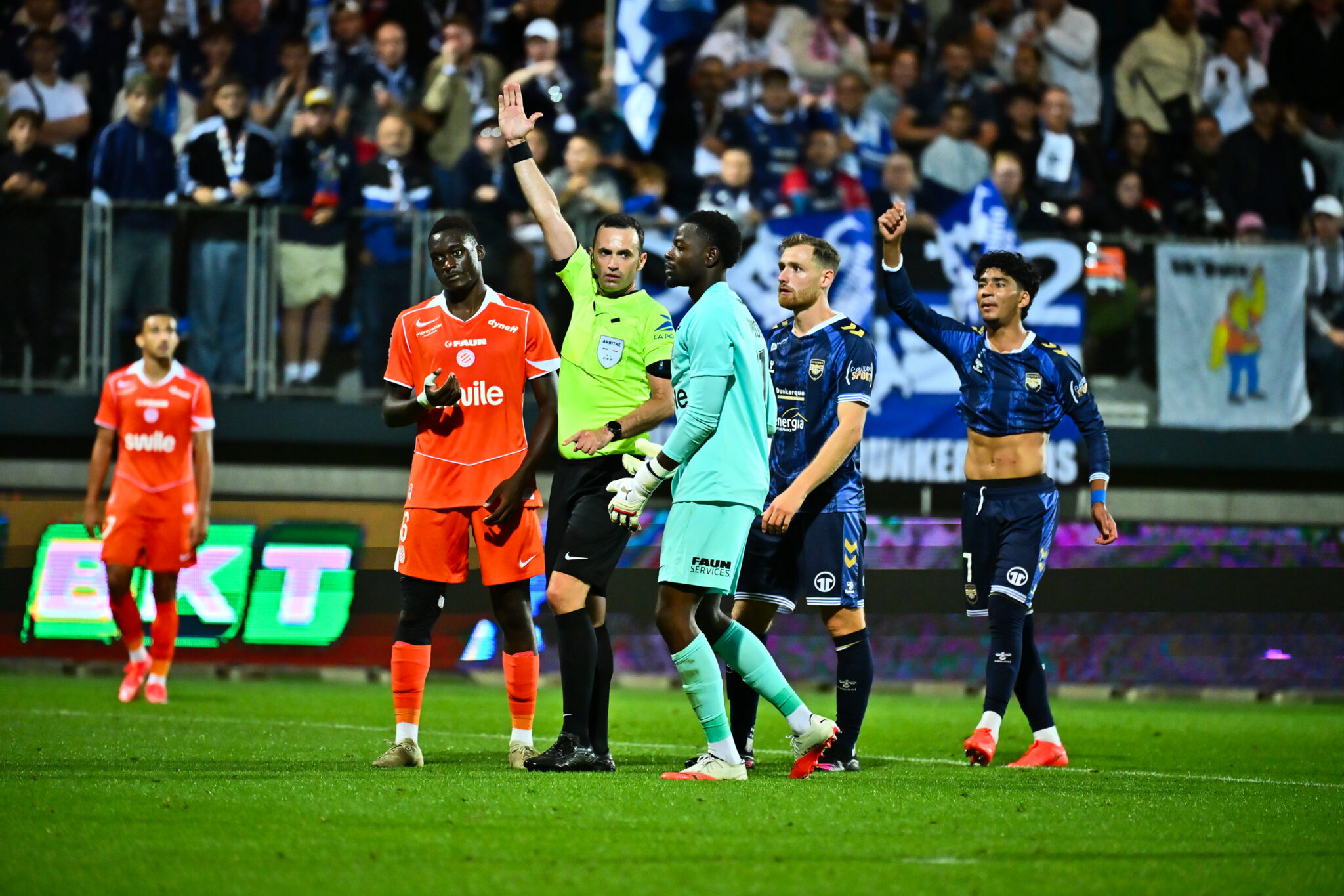 Soccer players on the field with the referee raising a hand, crowd watching from the stands behind them.