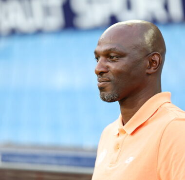 Profile view of a man in a peach polo shirt standing at a sports stadium, looking to the right.