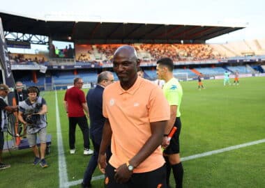 Soccer player in a peach polo walks along the sideline as camera crew and officials stand nearby on the field side of a stadium.