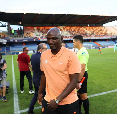 Soccer player in a peach polo walks along the sideline as camera crew and officials stand nearby on the field side of a stadium.