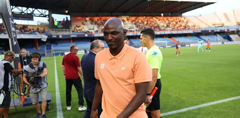 Soccer player in a peach polo walks along the sideline as camera crew and officials stand nearby on the field side of a stadium.