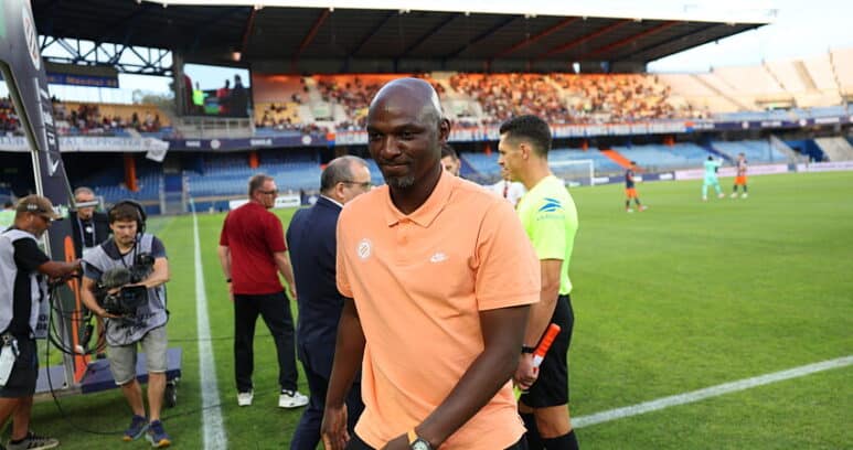 Soccer player in a peach polo walks along the sideline as camera crew and officials stand nearby on the field side of a stadium.