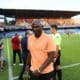 Soccer player in a peach polo walks along the sideline as camera crew and officials stand nearby on the field side of a stadium.