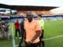 Soccer player in a peach polo walks along the sideline as camera crew and officials stand nearby on the field side of a stadium.