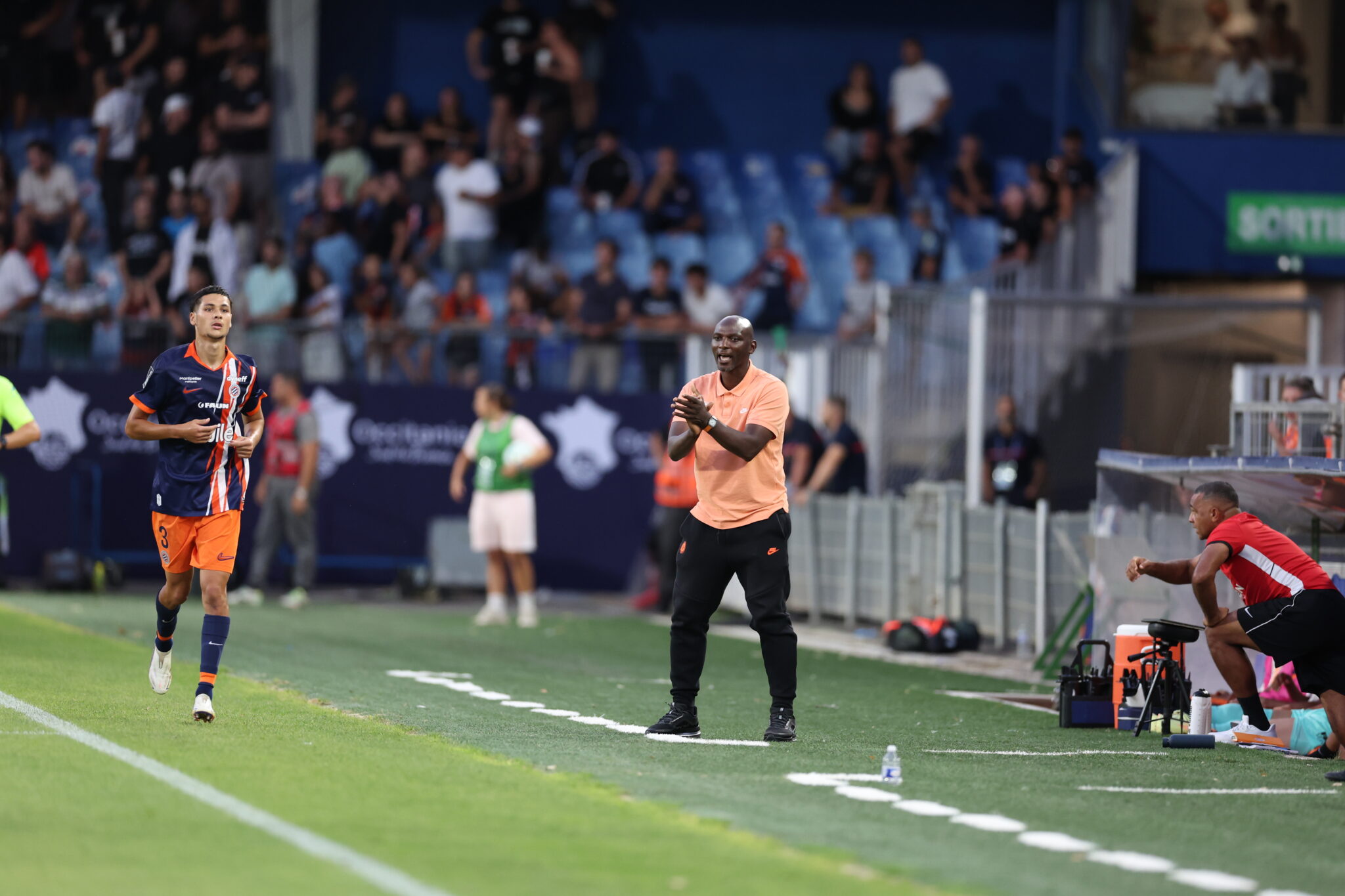 Naoufel El HANNACH Zoumana CAMARA Photo by Icon Sport - Photo by Icon Sport | AllezPaillade.com Soccer coach in a peach shirt claps on the sideline as a player in navy and orange runs by; blurred crowd behind.