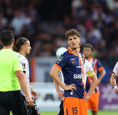 Soccer players on the field during a match; central player in navy/orange jersey (no. 15) looks to the side, surrounded by teammates and opponents, with a neon-green referee nearby and a crowd in the background.