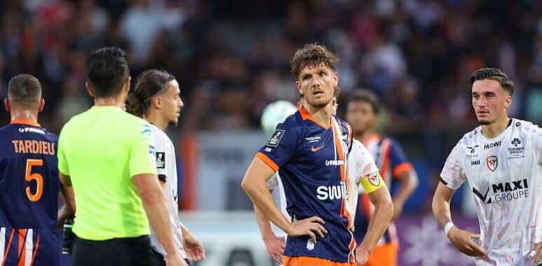 Soccer players on the field during a match; central player in navy/orange jersey (no. 15) looks to the side, surrounded by teammates and opponents, with a neon-green referee nearby and a crowd in the background.