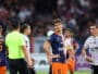 Soccer players on the field during a match; central player in navy/orange jersey (no. 15) looks to the side, surrounded by teammates and opponents, with a neon-green referee nearby and a crowd in the background.