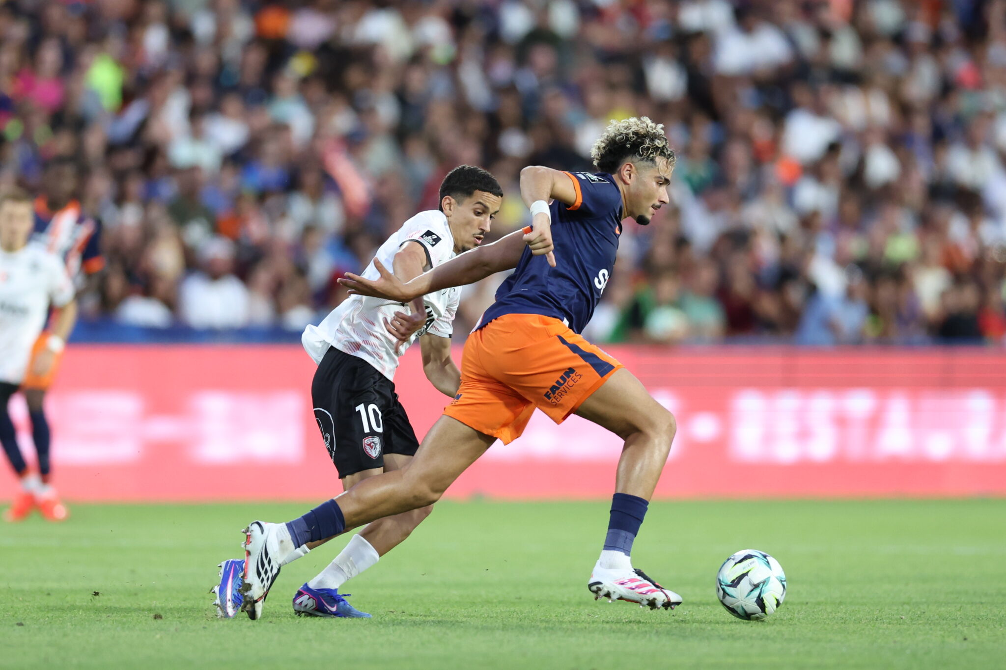 Daylam MEDDAH Photo by Icon Sport - Photo by Icon Sport | AllezPaillade.com Two soccer players chase the ball during a match, defender in white behind attacker in navy and orange as they sprint on the green field.