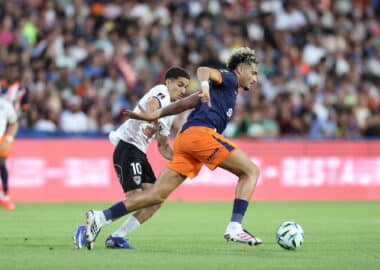 Two soccer players chase the ball during a match, defender in white behind attacker in navy and orange as they sprint on the green field.