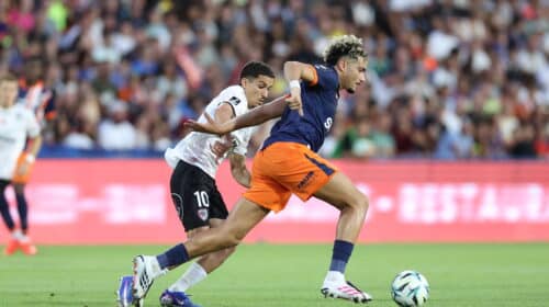 Two soccer players chase the ball during a match, defender in white behind attacker in navy and orange as they sprint on the green field.