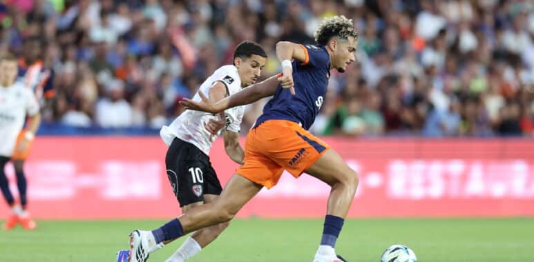 Two soccer players chase the ball during a match, defender in white behind attacker in navy and orange as they sprint on the green field.