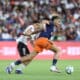 Two soccer players chase the ball during a match, defender in white behind attacker in navy and orange as they sprint on the green field.