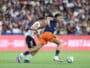 Two soccer players chase the ball during a match, defender in white behind attacker in navy and orange as they sprint on the green field.