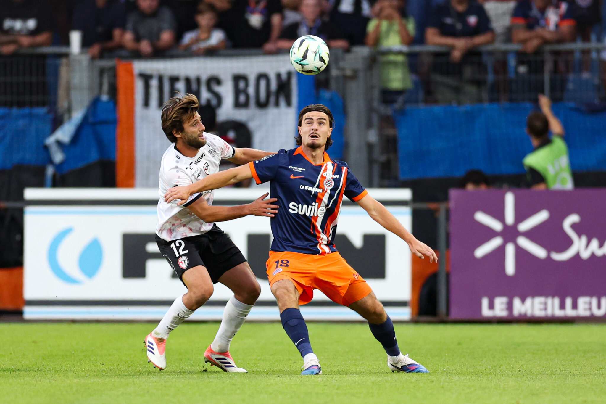 Two soccer players battle for a midair ball during a match, one in white and the other in navy and orange, with a cheering crowd behind them.