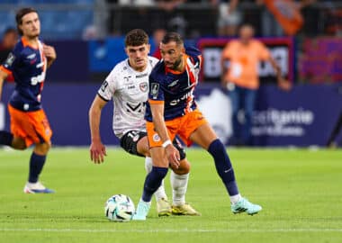 Soccer players in blue-orange and white chase the ball during a match on a sunny field.