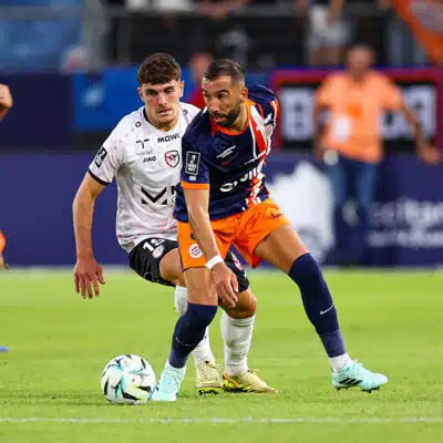 Soccer players in blue-orange and white chase the ball during a match on a sunny field.