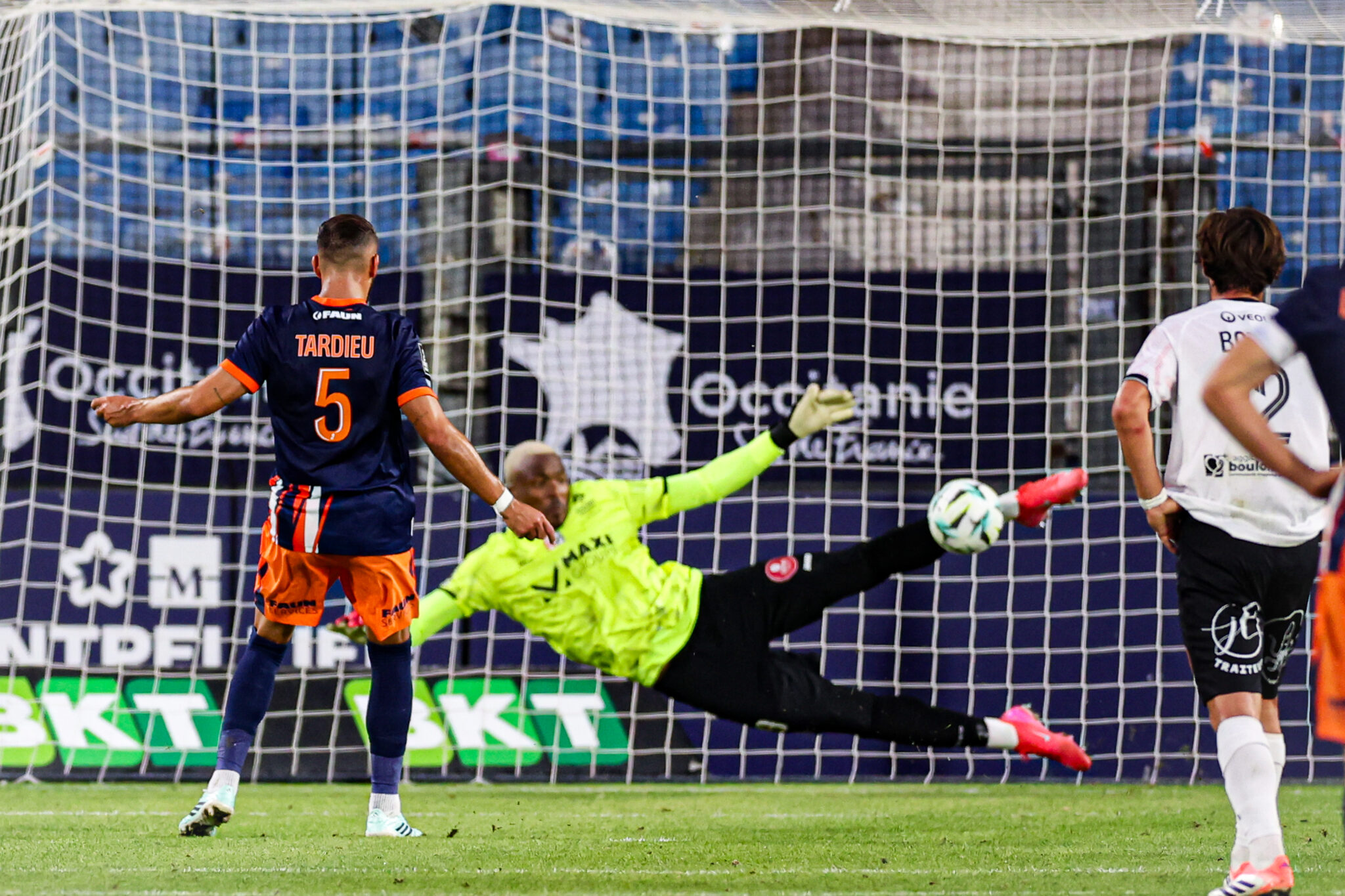 Montpellier Herault SC v US Boulogne - Ligue 2 BKT 20262027 | AllezPaillade.com Soccer scene with a goalkeeper in neon diving to stop a ball as two players watch near the goal net.