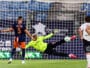 Soccer scene: goalkeeper in neon green dives to stop a shot as players watch near the goal net.