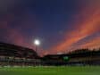 Soccer stadium at dusk with orange clouds, bright floodlights, and players on a green pitch.